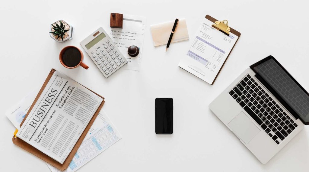 A busy desk with newspaper, coffee, a phone and a laptop.